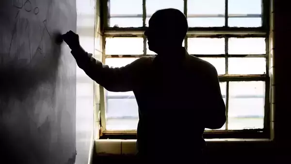 A man stands in profile writing on a chalkboard. He is silhouetted against a bright window.