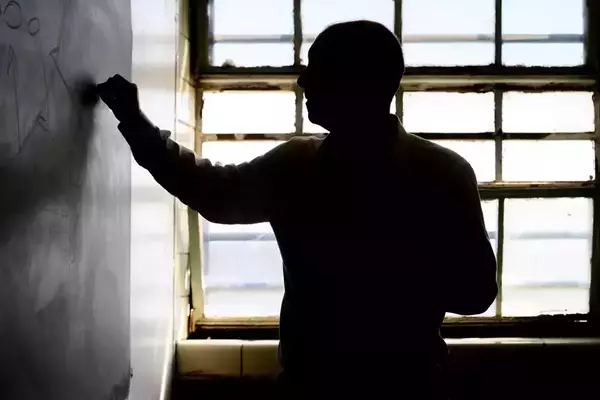 A man stands in profile writing on a chalkboard. He is silhouetted against a bright window.